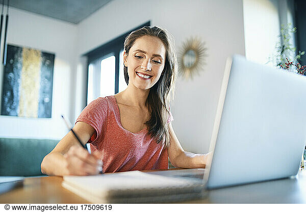 Smiling female freelancer writing in note pad on desk at home office