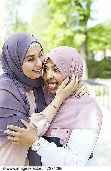 Smiling female friends looking away while hugging each other in public park