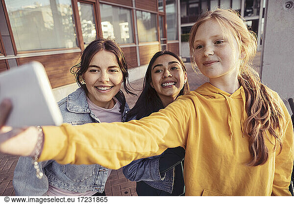 Smiling female friends taking selfie through smart phone on footpath