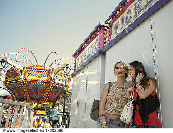 Smiling female friends talking while standing against ticket counter at amusement park