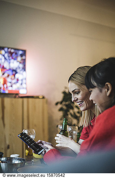 Smiling female friends using smart phone while watching soccer at home