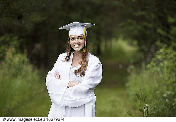 Smiling Female Graduate Wearing White Cap and Gown on Wooded Path