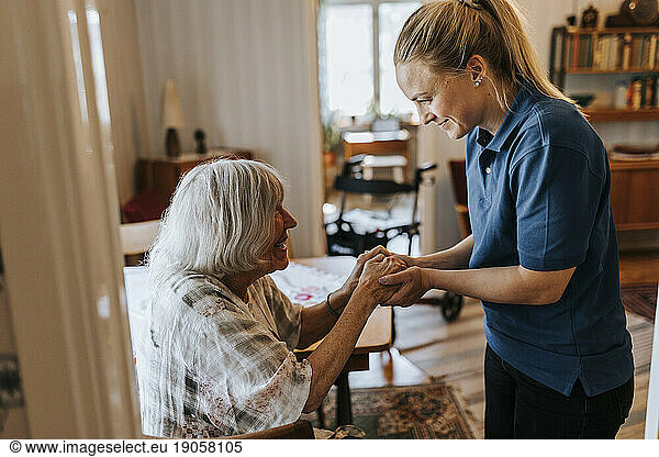 Smiling female healthcare worker holding hands of senior woman at home