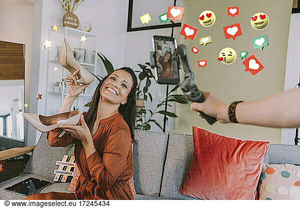 Smiling female influencer showing high heels while filming by friend through mobile phone by social icons at home