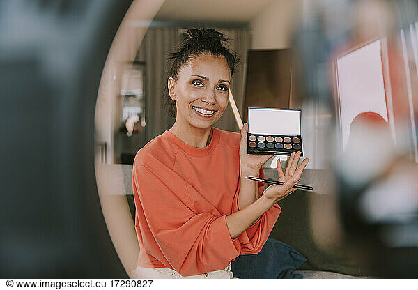 Smiling female influencer showing make-up palette while doing tutorial at home