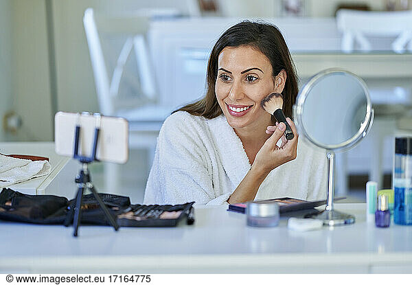 Smiling female influencer vlogging about make-up on smart phone at home