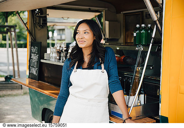 Smiling female owner looking away while standing against food truck