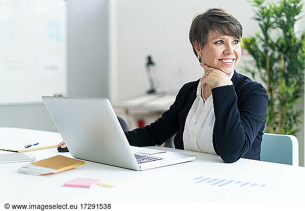 Smiling female professional sitting with laptop in office looking away