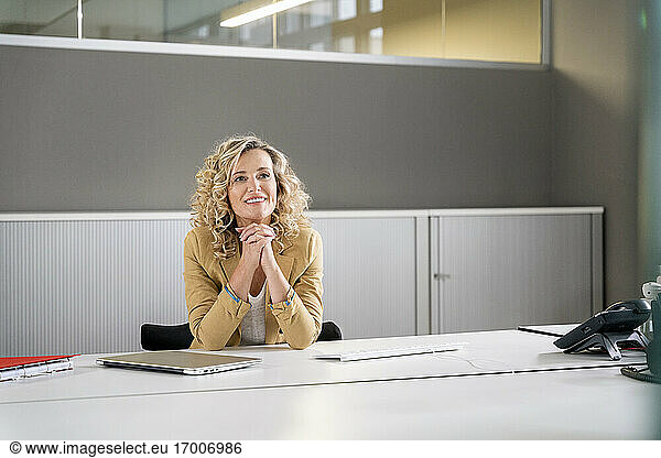 Smiling female professional with hands clasped day dreaming at desk in office