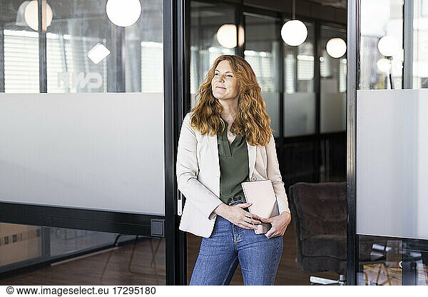 Smiling female professional with laptop leaning at doorway in office