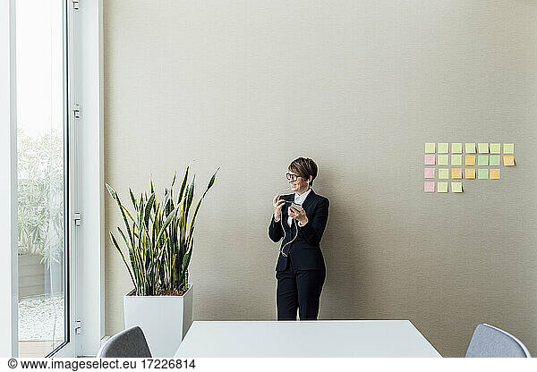 Smiling female professional with mobile phone looking away while standing against wall in office
