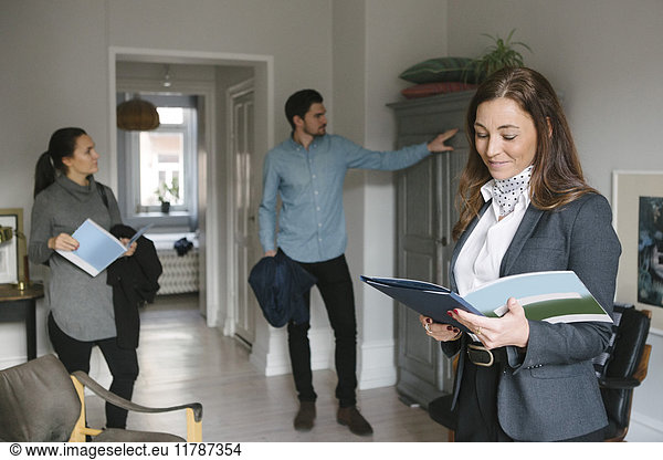 Smiling female real estate agent reading brochure while couple looking at cabinet at home