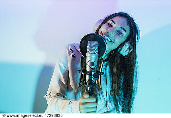 Smiling female singer wearing headphones singing in front of wall at studio