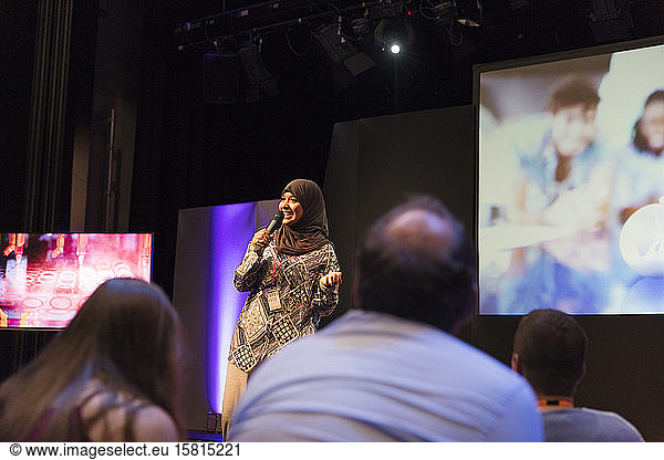 Smiling female speaker in hijab talking with microphone on stage