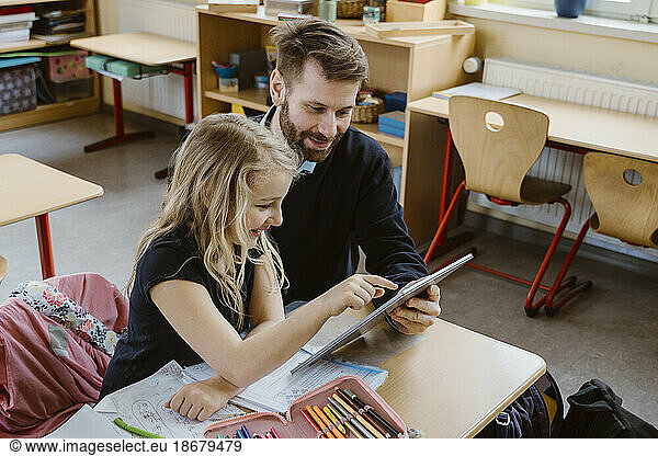 Smiling female student using tablet PC held by male teacher in classroom