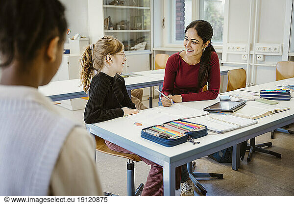 Smiling female teacher and schoolgirl interacting with each other while sitting in classroom