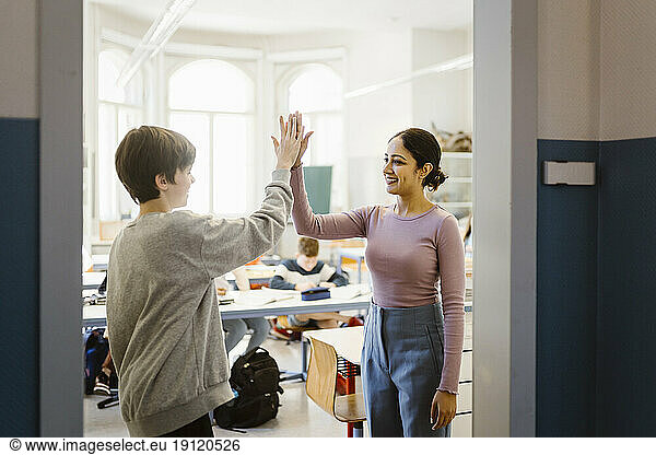 Smiling female teacher giving high-five to schoolboy standing in classroom