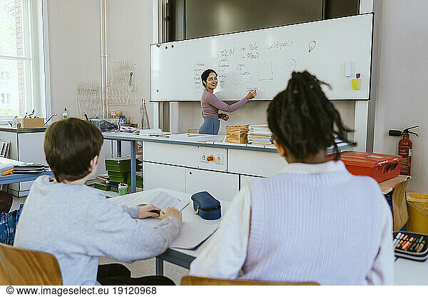 Smiling female teacher writing on whiteboard while teaching male students sitting in classroom