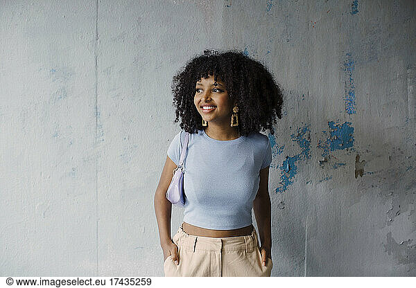 Smiling female teenager with hands in pockets against gray wall