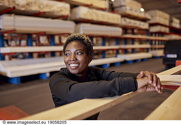 Smiling female worker looking away while leaning on plank in lumber industry