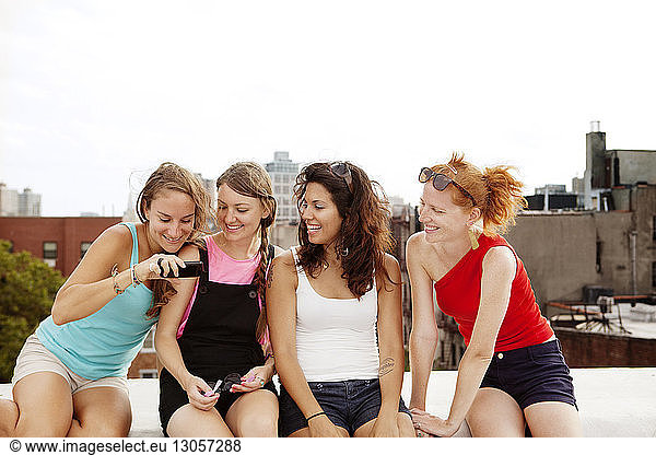 Smiling females looking at mobile phone while sitting on building terrace against clear sky