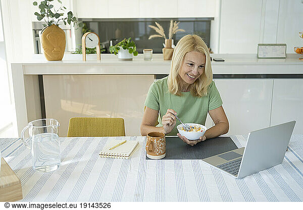 Smiling freelancer holding breakfast bowl in front of laptop at home