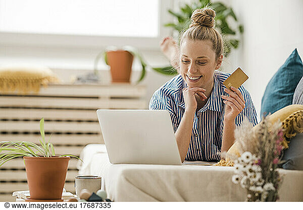 Smiling freelancer holding credit card looking at laptop lying on bed at home