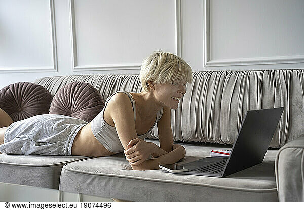 Smiling freelancer looking at laptop lying on sofa at home