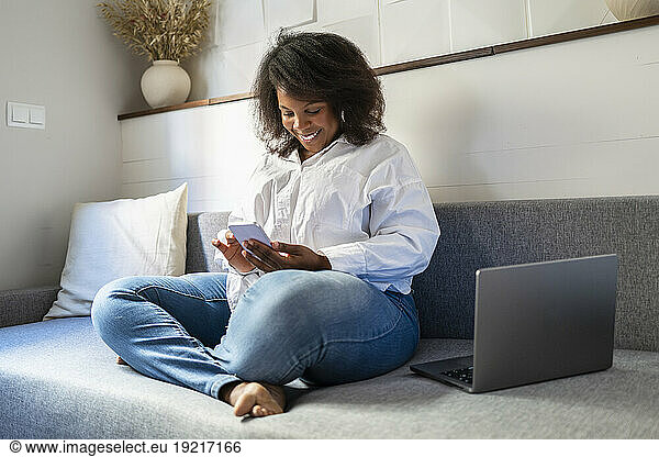Smiling freelancer sitting with laptop and using smart phone on sofa at home