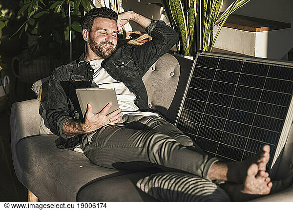 Smiling freelancer sitting with tablet PC and solar panel on sofa