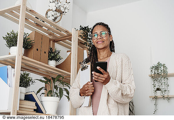 Smiling freelancer standing with clipboard and smart phone by shelf at home office