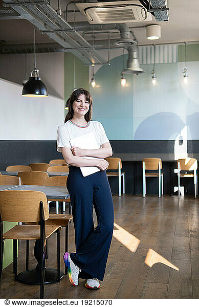 Smiling freelancer standing with laptop near table in cafe
