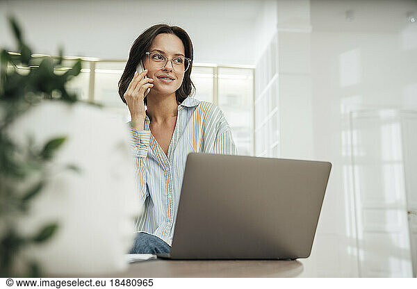 Smiling freelancer talking on smart phone and sitting with laptop at desk