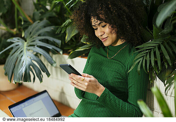Smiling freelancer text messaging through smart phone surrounded by plants