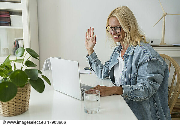 Smiling freelancer waving hand on video call through laptop at home