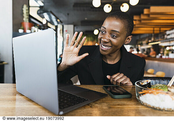 Smiling freelancer waving on video call over laptop in cafe