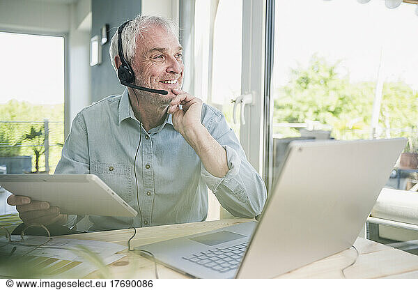Smiling freelancer wearing headset sitting with tablet PC and laptop at home