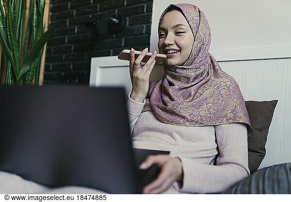 Smiling freelancer wearing hijab talking on speaker of phone at home