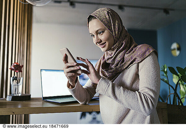 Smiling freelancer wearing hijab using smart phone in home office