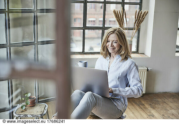 Smiling freelancer with coffee cup working on laptop at home