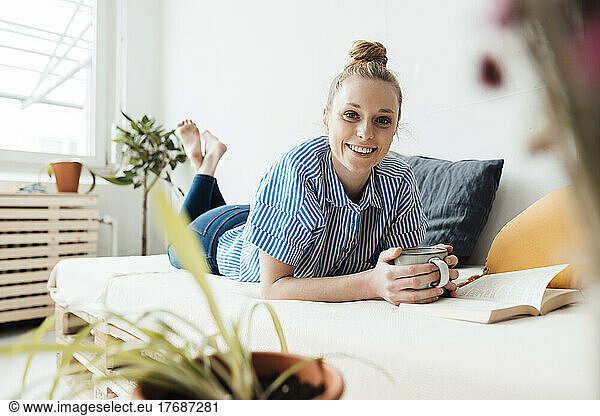 Smiling freelancer with coffee mug by book lying on bed at home