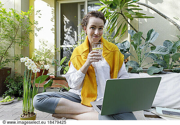 Smiling freelancer with laptop drinking water in backyard