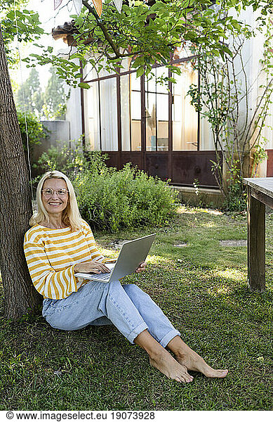 Smiling freelancer with laptop sitting on grass in garden