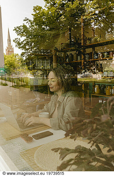 Smiling freelancer working on laptop in cafe seen through glass window