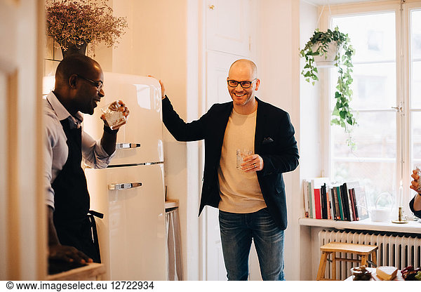 Smiling friends drinking water while standing by refrigerator in kitchen