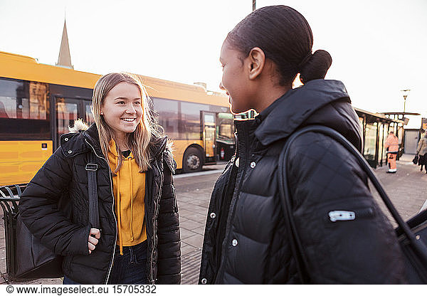 Smiling friends talking while standing against bus in city