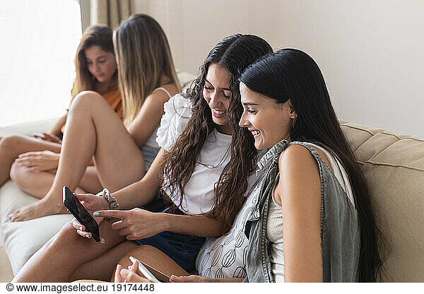 Smiling friends using smart phones sitting on sofa at home