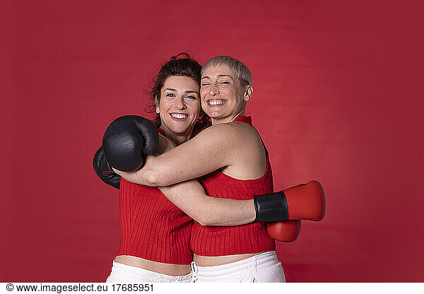 Smiling friends wearing boxing gloves hugging each other against red background
