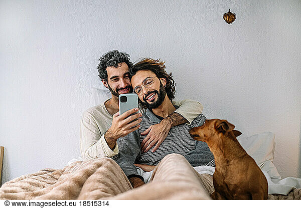 Smiling gay couple watching smart phone sitting on bed at home