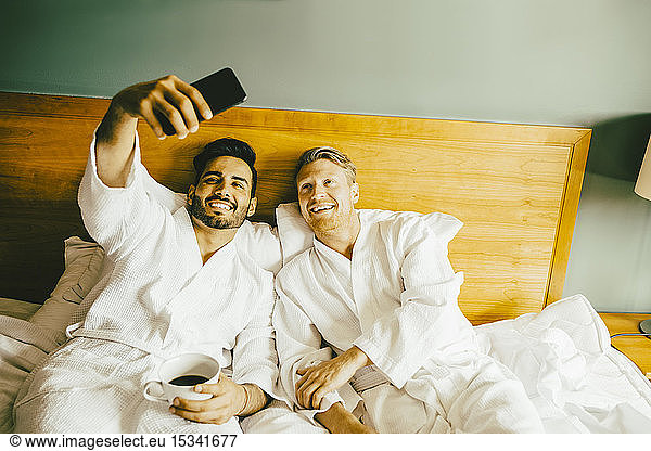 Smiling gay man holding coffee cup while taking selfie with boyfriend at hotel room